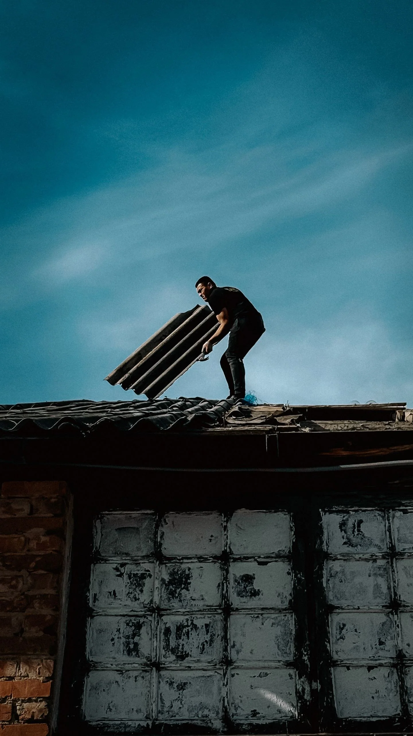 A man on a rooftop repairing with roofing sheets against a deep blue sky.